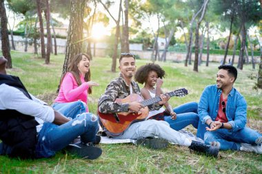 Çok ırklı arkadaşlar parkta gitar çalarken yerde oturup gülüyorlar. Arkadaşlık kavramı.