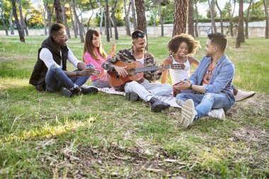 Biri parkta gitar çalarken, yerde oturan çok ırklı arkadaşlar. Arkadaşlık kavramı.