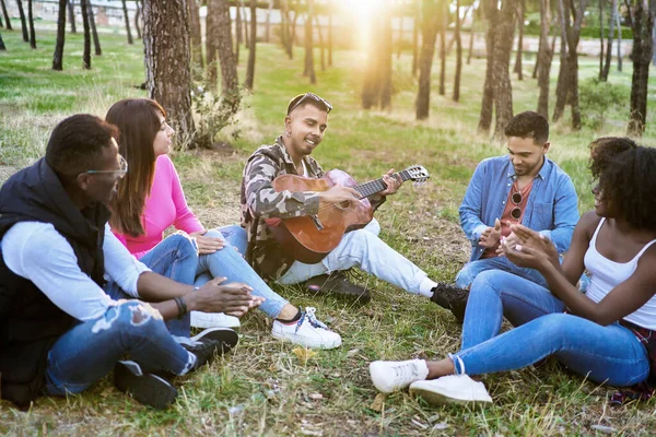 Biri parkta gitar çalarken, yerde oturan çok ırklı arkadaşlar. Arkadaşlık kavramı.