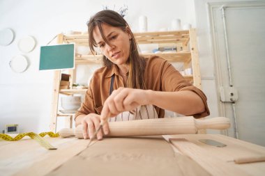 Woman in pottery workshop cutting clay while holding a rolling pin. Handicraft workshop 