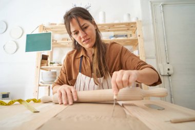 Woman in pottery workshop cutting clay while holding a rolling pin. Handicraft workshop 