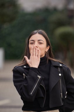 Stylish woman looking at camera and sending kiss while standing outdoors.