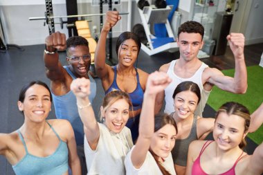 Çok kültürlü fitness grubu kameraya bakıp gülümsüyor ve yumruğunu havaya kaldırıyor.