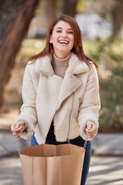 Bir kadın dışarıda alışveriş torbası tutarken kameraya bakıyor ve gülümsüyor. Alışveriş konsepti.