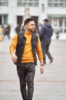 Latin man walking through the city streets wearing orange sweater and black vest. 