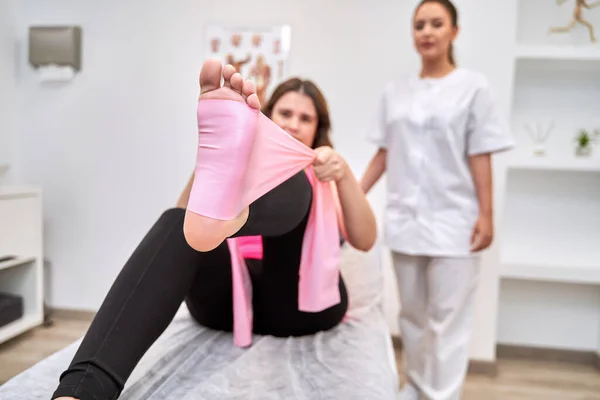 Experienced Physiotherapist Carefully Observes Female Patient Performing Leg Exercises with Elastic Bands