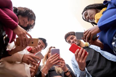 Çemberin içinde dikilirken cep telefonlarını kullanan çok ırklı arkadaşların düşük açılı görüntüsü. Arkadaşlık ve teknoloji kavramı.