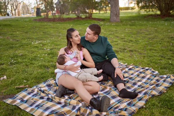 Jovem Casal Sentado Grama Parque Enquanto Alimenta Seu Bebê Olhando — Fotografia de Stock