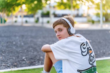 Genç bir çocuk dışarıda parkta kulaklıkla müzik dinlerken kameraya bakıyor. Teknoloji konsepti.