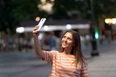 Geceleri sokakta cep telefonuyla selfie çekerken gülümseyen bir kadın. Teknoloji konsepti.