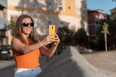 Güneş gözlüklü bir kadın güneşli bir günde dışarıda cep telefonuyla fotoğraf çekiyor. Teknoloji konsepti.