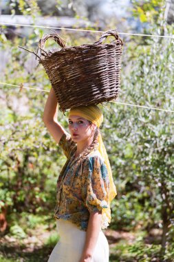 Çiftçi kadın bahçede kafasında hasır bir sepet taşırken kameraya bakıyor..