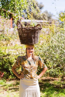 Kadın çiftçi kameraya bakıyor ve gülümsüyor. Bahçede kafasında hasır bir sepetle poz veriyor..