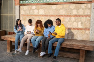 Etnik çeşitlilikte beş genç cep telefonlarına yoğunlaşıp, ahşap bir bankta oturuyorlar.