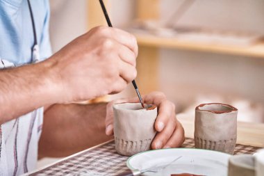 Bir çömlekçinin eli titizlikle el yapımı seramik bardakların kenarlarını ince bir fırçayla boyuyor.
