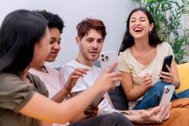 Bir grup neşeli arkadaş rahat bir kanepede gülüyor ve akıllı telefon ekranlarını paylaşıyorlar.. 