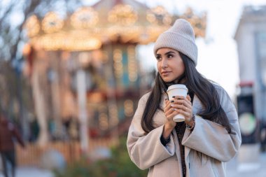 Yansıtıcı bir kadın, bir kış akşamında parıldayan atlıkarınca ışıklarının ortasındaki kahvesinden zevk alır.. 