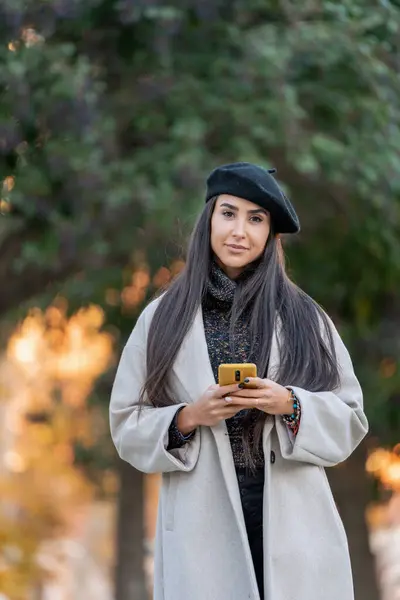 Chic woman in a beret using her smartphone in a park with autumn leaves in the background.