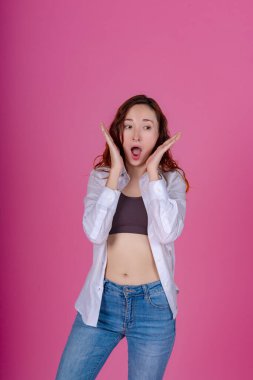 Şok olmuş genç bir kadın, elleri çerçeveli ve ağzı açık pembe bir zemine karşı şaşkınlık içinde.. 