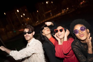 Bir grup arkadaş gece eğleniyorlar, güneş gözlüğü takıyorlar ve şakacı hareketler yapıyorlar.