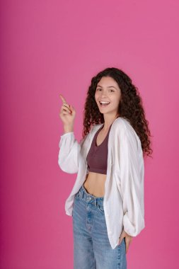 Beyaz hırkalı kıvırcık saçlı genç bir kadın ve yukarı bakan bir kot pantolon, neşeli bir gülümseme, pembe bir arka plan. 