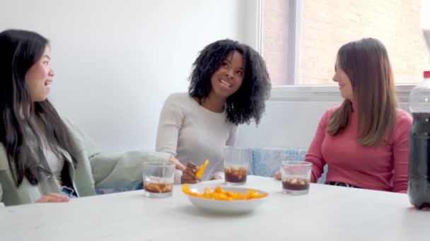 3人の多様な女性が カジュアルなソーシャル集会で明るくモダンなテーブルで 飲み物や軽食について笑いを共有しています — ストック動画