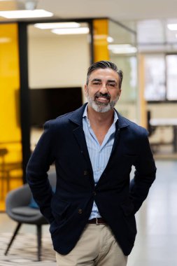 Sakallı, spor ceket giyen, aydınlık bir ofis koridorunda poz veren neşeli, olgun bir adam.. 