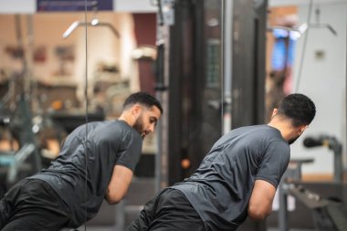 Beden eğitiminde arka eklenti makinesi kullanan konsantre bir adam, ayna yansıtma çabası ile. 