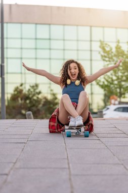 Şen şakrak genç bir kadın, kulaklıklarıyla kaykayın üzerinde oturuyor, kollarını kaldırıp kutlama yapıyor, şehir parkında mavi bir atlet ve ekose gömlek giyiyor.. 