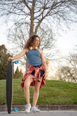 Kıvırcık saçlı genç bir kadın kaykay ve kulaklıkla güvenle ayakta duruyor. Gün batımında parkta mavi bir atlet ve ekose gömlek giyiyor.. 
