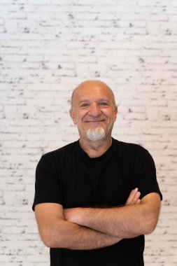 Siyah gömlekli gülümseyen yaşlı bir adam kollarını kavuşturmuş, beyaz tuğla bir duvara yaslanmış, kendinden emin ve rahat görünüyor.. 