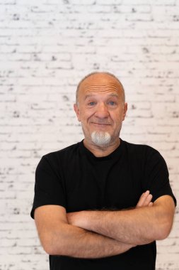 Çapraz kollu ve siyah gömlekli yaşlı bir adam, beyaz tuğladan bir arka plan karşısında duruyor, kameraya bakıyor.. 