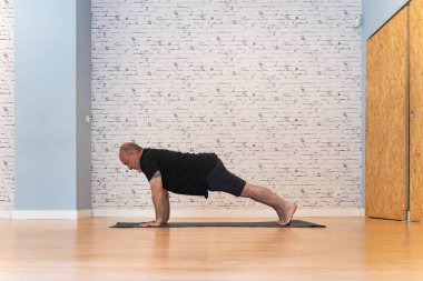 Siyah kıyafetli son sınıf öğrencisi elinde kalas yoga pozuyla paspasın üzerinde beyaz tuğladan bir duvar ve ahşap zemin olan ferah bir odada duruyor.. 