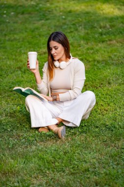 Beyaz elbiseli bir kadın kahve fincanı tutuyor, kitap okuyor ve parkta çimlerin üzerinde oturuyor.. 