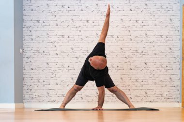 Siyah kıyafetli son sınıf öğrencisi, beyaz tuğla duvarlı ve ahşap zeminli bir odada paspasın üzerinde üçgen yoga pozu veriyor.. 