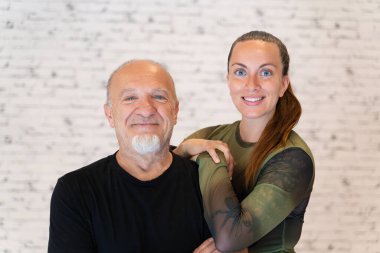 Siyah gömlekli gülümseyen yaşlı bir adam ve saf yeşil bluzlu genç bir kadın kollarını birbirine dolamış, beyaz tuğla bir arka plana yaslanmış.. 