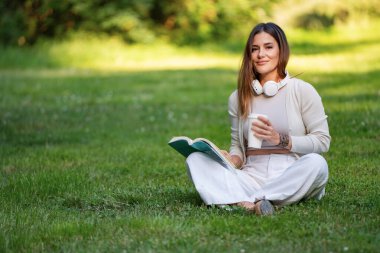 Kulaklık takan, elinde kahve fincanı tutan ve parkta çimenlerde otururken kitap okuyan bir kadın.. 