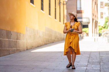 Sarı elbiseli ve şapkalı bir kadın elinde kamerayla güneşli bir sokakta yürüyor. Aydınlık bir günde şehri gezen bir turist. 