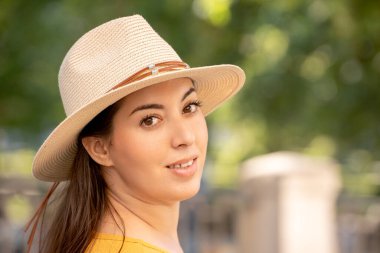 Güneş şapkası takan bir kadının yakın çekimi, yumuşak bir gülümsemeyle geriye bakıyor. Güneşli bir günde dışarıda, sakin ve sakin bir tavır sergiliyor.. 