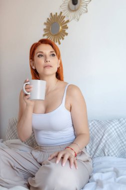 Kızıl saçlı kadın bir yatağa oturur, elinde bir kahve fincanı tutarak düşünceli bir şekilde bakar. Yakınlarda açık bir kitap var ve üzerinde beyaz bir atlet ve bej pantolon var.