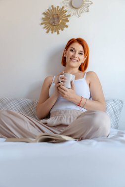 Kızıl saçlı kadın elinde kahve fincanı tutarken sıcak bir şekilde gülümsüyor, açık bir kitapla yatakta rahatça oturuyor..