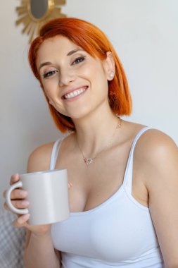 Elinde kahve fincanı tutarken parlak bir şekilde gülümseyen kızıl saçlı bir kadının yakın çekimi. Beyaz bir atlet giyiyordu ve kalp şeklinde bir kolye takıyordu. 
