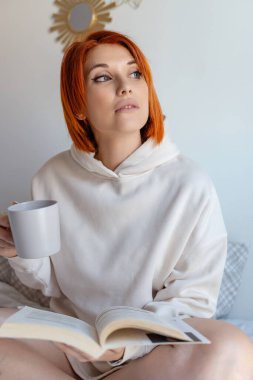 Bej kapüşonlu, kızıl saçlı bir kadın elinde kahve fincanıyla yatağında oturmuş kitap okurken dikkatlice seyrediyor..