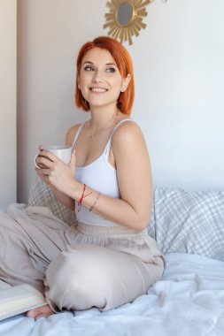 Kızıl saçlı kadın kahve fincanı tutarken sıcak bir şekilde gülümsüyor, yanında açık bir kitapla rahatça bir yatakta oturuyor.. 