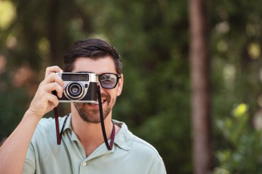 Bir adam güneşli bir parkta eski bir kamerayla fotoğraf çekerken gülümsüyor. Açık havayı sever, yeşilliklerle çevrili, rahat ve neşeli bir anda.. 