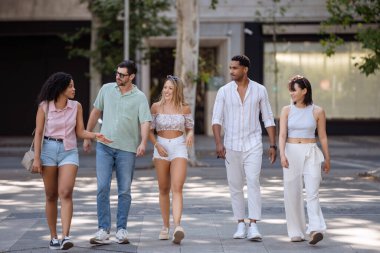 Şehir meydanında gezen beş genç arkadaştan oluşan çeşitli bir grup ağaçlar ve kentsel çevreyle güneşli bir günün tadını çıkarıyorlar.. 