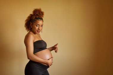 Hamile bir kadın kameraya bakıyor ve göbeğini gösterip izole edilmiş arka planda duruyor. Hamilelik ve annelik kavramı.