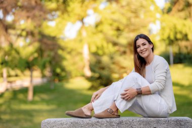 Güzel bir kadın, huzurlu bir park ortamında, taş bir bankta dinlenirken gülümsüyor.