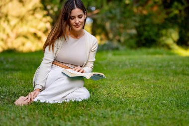Genç bayan parkta huzurlu bir öğleden sonranın tadını çıkarıyor, çimlerin üzerinde oturuyor ve kitap okuyor.