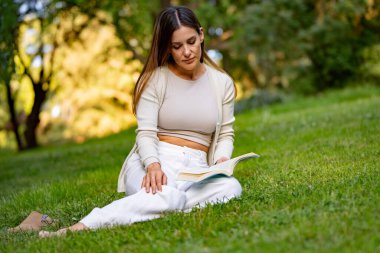 Genç bir kadın, güneşli bir günde bir kitap okumaya dalmış, parkta çimlerin üzerinde oturuyor.